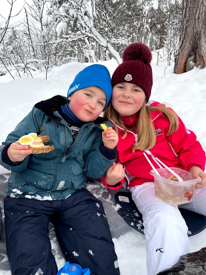 Ole og Nicoline sitter i snøen i skiklær og spiser matpakke.